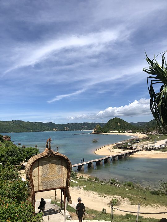 A bamboo footbridge crossing to a curved beach below green hills, seen through a thatched gateway